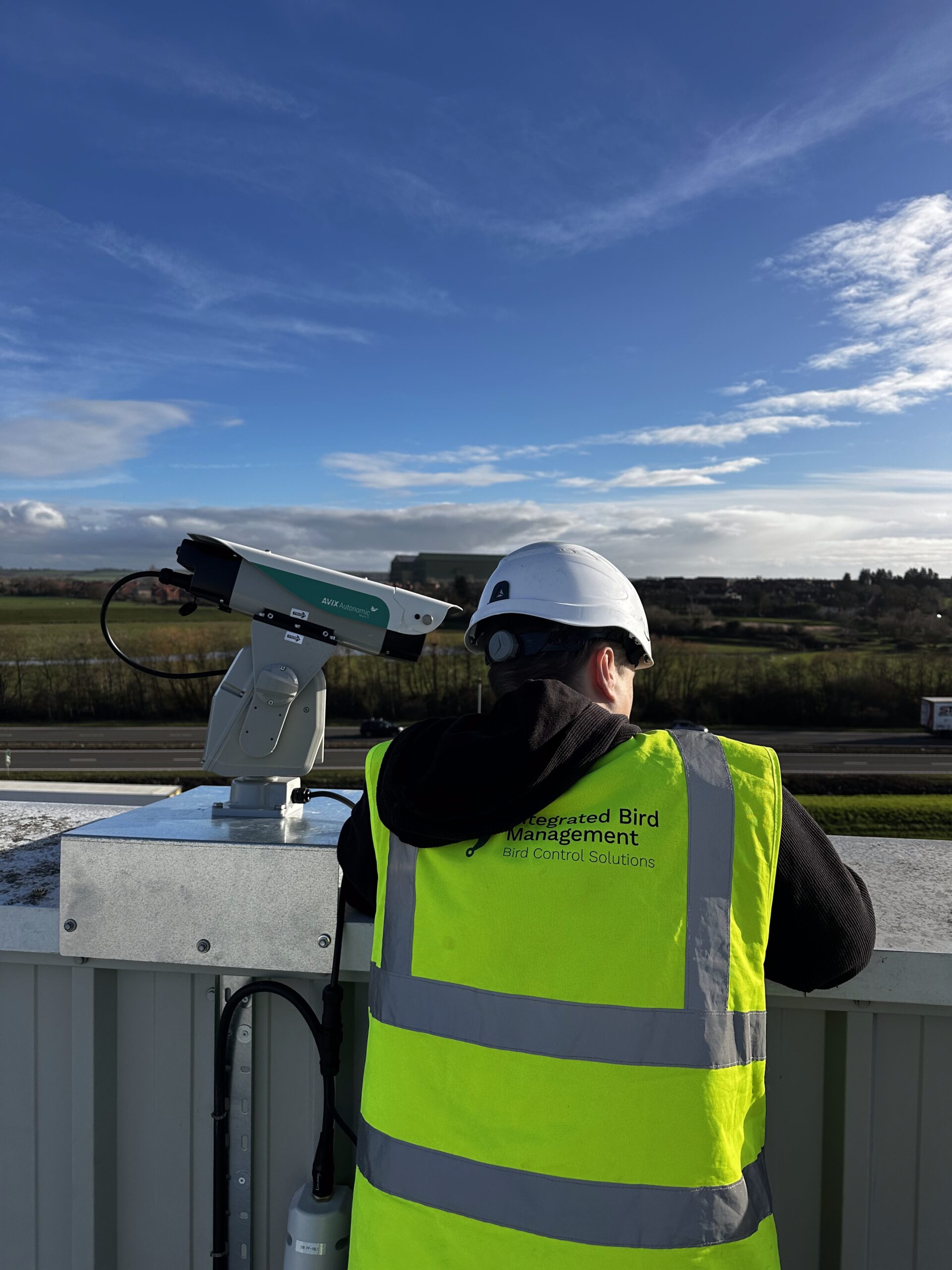 Integrated Bird Management technician installing Avix Autonomic Mark II laser bird deterrent on rooftop of distribution building, Bedford