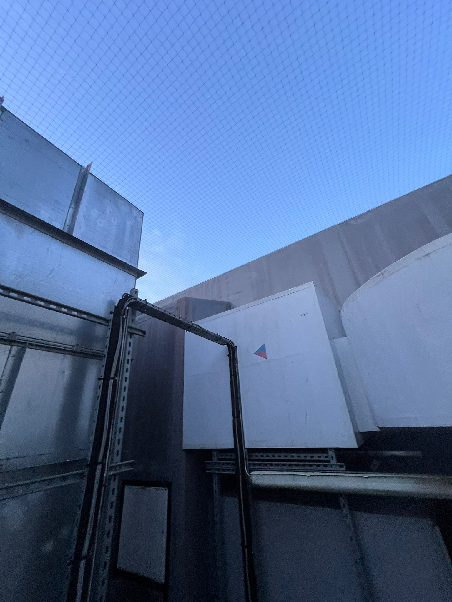 View upward within a narrow service area showing metal ducting, electrical conduits and equipment units, with protective netting installed overhead.