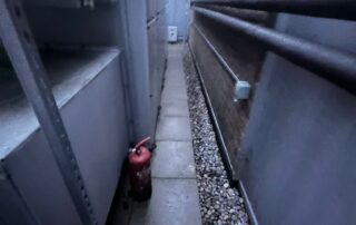 Narrow outdoor service corridor between building walls with paving slabs, a gravel drainage strip, exposed pipes on the right wall, and a red fire extinguisher placed on the ground.